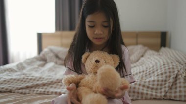 Tatlı Asyalı küçük kız evinde yatağında otururken oyuncak ayıya sarılıyor ve gülümsüyor.