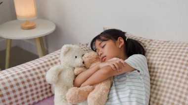 Asyalı kız, oyuncak ayılara sarılırken, rahat bir yatak odasında masumiyeti, güvenliği ve sıcaklığı temsil ederken, huzurlu bir şekilde yatakta uyuyor..