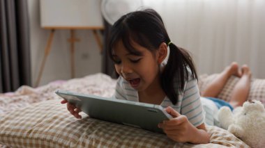 Rahat yatak odasında tablet kullanan odaklanmış Asyalı kız, sıcak ve güvenli bir ev ortamında modern dijital öğrenme ve ekran zamanını temsil ediyor.