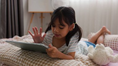 Rahat yatak odasında tablet kullanan odaklanmış Asyalı kız, sıcak ve güvenli bir ev ortamında modern dijital öğrenme ve ekran zamanını temsil ediyor.