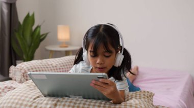 Kulaklık takan ve yatakta yatarken tablet kullanan Asyalı kız, evde dijital öğrenme veya eğlenceye olan ilgisini ve eğlencesini ifade ediyor.