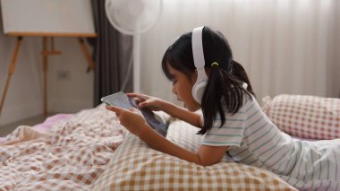 Kulaklık takan ve yatakta yatarken tablet kullanan Asyalı kız, evde dijital öğrenme veya eğlenceye olan ilgisini ve eğlencesini ifade ediyor.