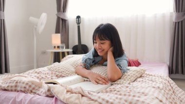 Asyalı kız kitap okurken gülümsüyor ve sıcak bir ortamda rahat ve mutlu bir şekilde kendini öğrenmeyi temsil eden rahat yatak odasında ders çalışıyor..