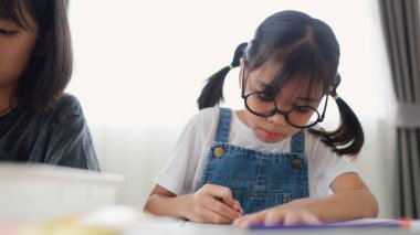 İki Asyalı kız, sıcak ve odaklı bir atmosferle evde eğitim, yaratıcılık ve modern eğitimi temsil ederek, parlak bir odada okuyor ve resim çiziyor..