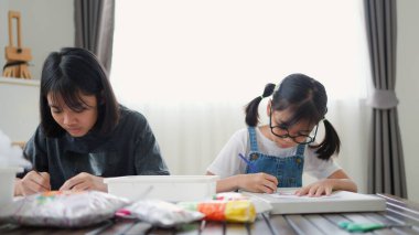 İki Asyalı kız, sıcak ve odaklı bir atmosferle evde eğitim, yaratıcılık ve modern eğitimi temsil ederek, parlak bir odada okuyor ve resim çiziyor..