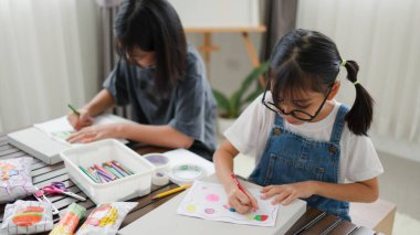 İki Asyalı kız, sıcak ve odaklı bir atmosferle evde eğitim, yaratıcılık ve modern eğitimi temsil ederek, parlak bir odada okuyor ve resim çiziyor..