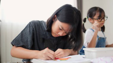 İki Asyalı kız, sıcak ve odaklı bir atmosferle evde eğitim, yaratıcılık ve modern eğitimi temsil ederek, parlak bir odada okuyor ve resim çiziyor..