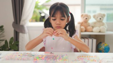 Asyalı kız evde renkli boncuklara iplik geçirip sakin bir ortamda konsantrasyonu, yaratıcılığı ve iyi motor becerileri arttıran eğlenceli ve eğitici bir aktiviteye odaklanıyor..