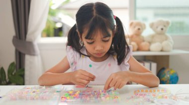 Asyalı kız evde renkli boncuklara iplik geçirip sakin bir ortamda konsantrasyonu, yaratıcılığı ve iyi motor becerileri arttıran eğlenceli ve eğitici bir aktiviteye odaklanıyor..