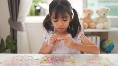 Asyalı kız evde renkli boncuklara iplik geçirip sakin bir ortamda konsantrasyonu, yaratıcılığı ve iyi motor becerileri arttıran eğlenceli ve eğitici bir aktiviteye odaklanıyor..