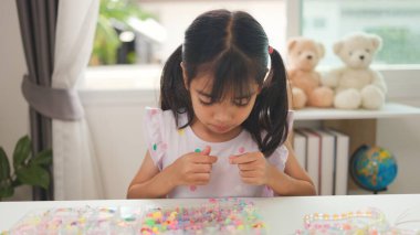 Asyalı kız evde renkli boncuklara iplik geçirip sakin bir ortamda konsantrasyonu, yaratıcılığı ve iyi motor becerileri arttıran eğlenceli ve eğitici bir aktiviteye odaklanıyor..