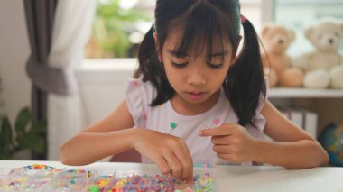 Asyalı kız evde renkli boncuklara iplik geçirip sakin bir ortamda konsantrasyonu, yaratıcılığı ve iyi motor becerileri arttıran eğlenceli ve eğitici bir aktiviteye odaklanıyor..