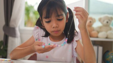 Asyalı kız evde renkli boncuklara iplik geçirip sakin bir ortamda konsantrasyonu, yaratıcılığı ve iyi motor becerileri arttıran eğlenceli ve eğitici bir aktiviteye odaklanıyor..