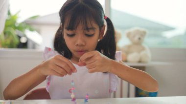 Asyalı kız evde renkli boncuklara iplik geçirip sakin bir ortamda konsantrasyonu, yaratıcılığı ve iyi motor becerileri arttıran eğlenceli ve eğitici bir aktiviteye odaklanıyor..