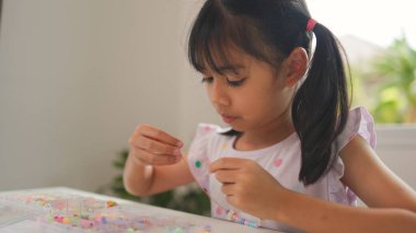 Asyalı kız evde renkli boncuklara iplik geçirip sakin bir ortamda konsantrasyonu, yaratıcılığı ve iyi motor becerileri arttıran eğlenceli ve eğitici bir aktiviteye odaklanıyor..