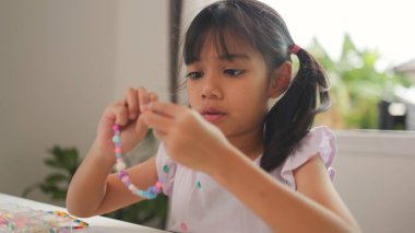 Asyalı kız evde renkli boncuklara iplik geçirip sakin bir ortamda konsantrasyonu, yaratıcılığı ve iyi motor becerileri arttıran eğlenceli ve eğitici bir aktiviteye odaklanıyor..