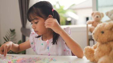 Asyalı kız evde renkli boncuklara iplik geçirip sakin bir ortamda konsantrasyonu, yaratıcılığı ve iyi motor becerileri arttıran eğlenceli ve eğitici bir aktiviteye odaklanıyor..