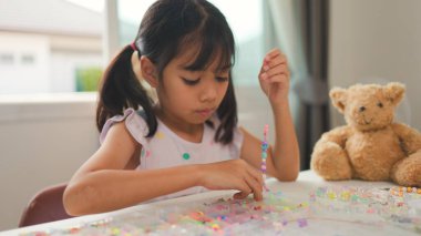 Asyalı kız evde renkli boncuklara iplik geçirip sakin bir ortamda konsantrasyonu, yaratıcılığı ve iyi motor becerileri arttıran eğlenceli ve eğitici bir aktiviteye odaklanıyor..