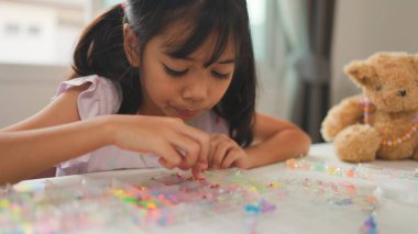 Asyalı kız evde renkli boncuklara iplik geçirip sakin bir ortamda konsantrasyonu, yaratıcılığı ve iyi motor becerileri arttıran eğlenceli ve eğitici bir aktiviteye odaklanıyor..