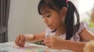 Asyalı kız evde renkli boncuklara iplik geçirip sakin bir ortamda konsantrasyonu, yaratıcılığı ve iyi motor becerileri arttıran eğlenceli ve eğitici bir aktiviteye odaklanıyor..