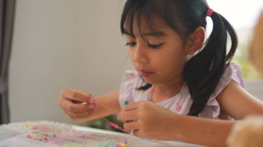 Asyalı kız evde renkli boncuklara iplik geçirip sakin bir ortamda konsantrasyonu, yaratıcılığı ve iyi motor becerileri arttıran eğlenceli ve eğitici bir aktiviteye odaklanıyor..