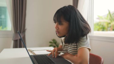 Asyalı kız evde odaklanmış bir şekilde okuyor, dizüstü bilgisayar kullanırken bir deftere yazıyor, evde eğitimi, uzaktan eğitimi ve üretken eğitimi sakin bir alanda temsil ediyor..