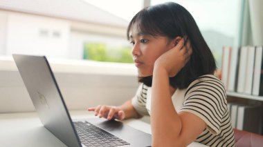 Asyalı genç kız evde dizüstü bilgisayarla online bir sınıfı dikkatlice izliyor, sakin bir çalışma alanında odaklanmış dijital öğrenimi, ev okulunu ve uzaktan eğitimi temsil ediyor..