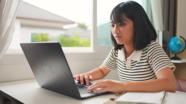 Evde online öğrenim için dizüstü bilgisayar kullanan Asyalı kız, sakin ve odaklı bir çalışma ortamında uzaktan eğitimi, ev okulunu ve dijital beceri geliştirmeyi temsil ediyor.