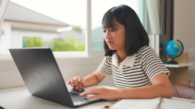 Evde online öğrenim için dizüstü bilgisayar kullanan Asyalı kız, sakin ve odaklı bir çalışma ortamında uzaktan eğitimi, ev okulunu ve dijital beceri geliştirmeyi temsil ediyor.