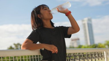 Açık hava egzersizlerinden sonra plastik şişeden su içen Asyalı genç kız, kentsel arka planda doğal güneş ışığı altında hidrasyon, spor ve sağlıklı yaşam tarzını teşvik ediyor..