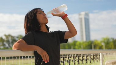 Açık hava egzersizlerinden sonra plastik şişeden su içen Asyalı genç kız, kentsel arka planda doğal güneş ışığı altında hidrasyon, spor ve sağlıklı yaşam tarzını teşvik ediyor..