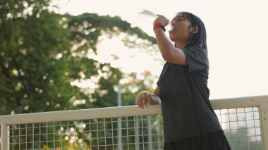 Açık hava egzersizlerinden sonra plastik şişeden su içen Asyalı genç kız, kentsel arka planda doğal güneş ışığı altında hidrasyon, spor ve sağlıklı yaşam tarzını teşvik ediyor..