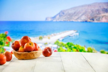Ahşap kasedeki şeftalinin taze meyvesi ahşap masa üstünde güzel bulanık deniz ve mavi güneşli gökyüzü arkaplanı. Bazıları uzaktaki Yunanistan binalarını bulanıklaştırdı.