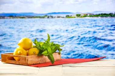 Masanın üstünde sarı limonlu ahşap tepsi ve arka planda güzel bir okyanus, dağlık kayalar ve adalar ve berrak gökyüzü..