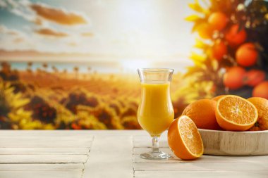Portakal meyvesi ve ahşap masanın üstünde cam içinde taze portakal suyu. Bahçedeki turunçgillerden oluşan güzel bulanık bir manzara. Tatil zamanı ve sıcak gün için soğuk bir içecek..