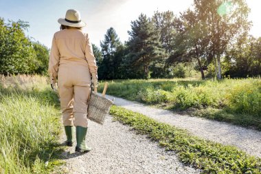 Bahçecilik tulumlu, şapkalı, çayırlarla tarlalar arasında yürüyen bir çiftçi kadın. Yeşillik, ağaçlar ve bitkiler. Güneşli sonbahar bahçıvanlığı.