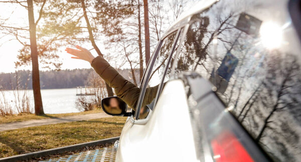 Закрыть женскую руку, машущую из окна машины. Вождение автомобиля на дороге в солнечный день.