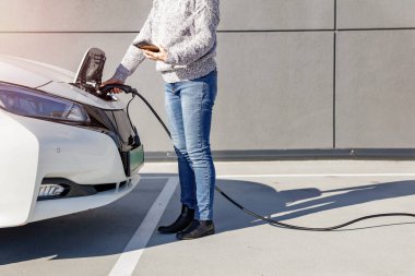 Akıllı telefonu olan bir kadın park yerindeki şarj kablosuna bağlı beyaz elektrikli bir arabanın yanında duruyor..