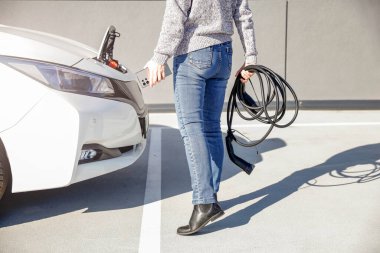 Beyaz elektrikli bir aracın yanında, sarmalanmış bir kablo tutan kazak ve kot giyen biri. Arabanın şarj bölmesi açık ve sahne açık bir gökyüzünün altındaki asfalt bir alanda çekiliyor.