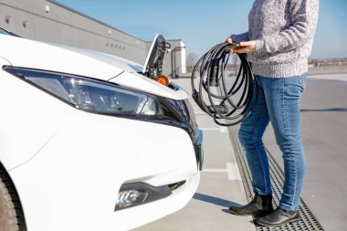 Beyaz elektrikli bir aracın yanında, sarmalanmış bir kablo tutan kazak ve kot giyen biri. Arabanın şarj bölmesi açık ve sahne açık bir gökyüzünün altındaki asfalt bir alanda çekiliyor.