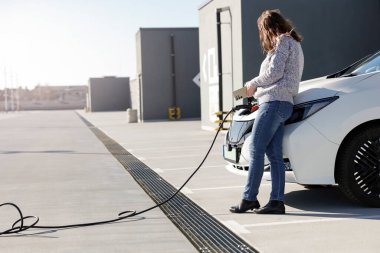 Akıllı telefonu olan bir kadın park yerindeki şarj kablosuna bağlı beyaz elektrikli bir arabanın yanında duruyor..