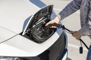 Kadın elektrik kablosunu elektrikli arabaya bağlıyor. Aracın şarj portu açık ve kişinin elinde siyah şarj prizi var..