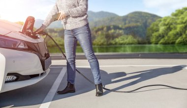 Elektrikli araba şarj istasyonu kullanan bir kadın. Sıradan kıyafetler giymiş bir kadın, beton otoparkta Ca 'nın yanında dururken cep telefonu kullanıyor. Bulanık göl ve dağ arkaplanı.