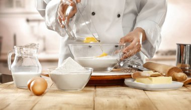 Ahşap mutfak tezgahında aşçı üniforması giyen biri büyük cam kaseye un karıştırarak yumurta döküyor. Mutfak içi arka plan.