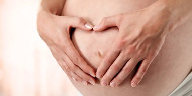 Elleri yavaşça hamile karnına yerleştirilir ve kalp şekli oluşur. Parlak bir arka plan. Anne sevgisini ifade eden fotoğraf.