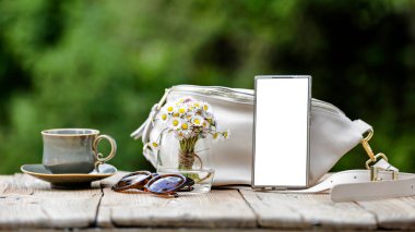 Beyaz el çantası, beyaz papatyalı cam vazo, boş ekranlı akıllı telefon, güneş gözlüğü ve çay tabağıyla kahve fincanı..