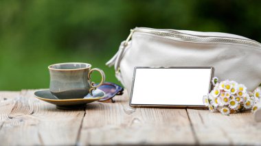 Beyaz el çantası, beyaz papatyalı cam vazo, boş ekranlı akıllı telefon, güneş gözlüğü ve çay tabağıyla kahve fincanı..