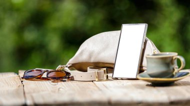 Bahçe masasının üzerinde fincan ve fincan tabağı, akıllı telefon, güneş gözlüğü ve bulanık bahçeye ayarlanmış el çantası ile bahar kompozisyonu. Anneler Günü ya da Kadınlar Günü ya da fotokopi alanı olan tebrik kartı geçmişi.