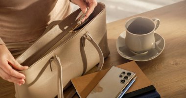 Bir kadının elleri masanın üzerine bir el çantasının fermuarını çekerken, iş günü için hazırlanmayı öneriyor..