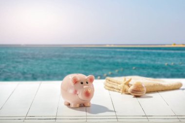 Kumsalın yanındaki Tahta Güverte Tatil Tasarruf Tasarruf Konsepti 'nde Piggy Bank ve Havlu. Boşluğu, bulanık sahili ve deniz arkaplanını kopyala.